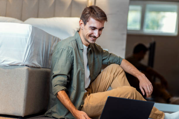 istock Man is sitting next to the bed in bedroom on the floor using a laptop. 2198643283