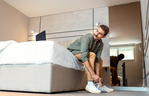 istock Man sitting on bed in bedroom putting on sneakers on while using laptop. 2198643423