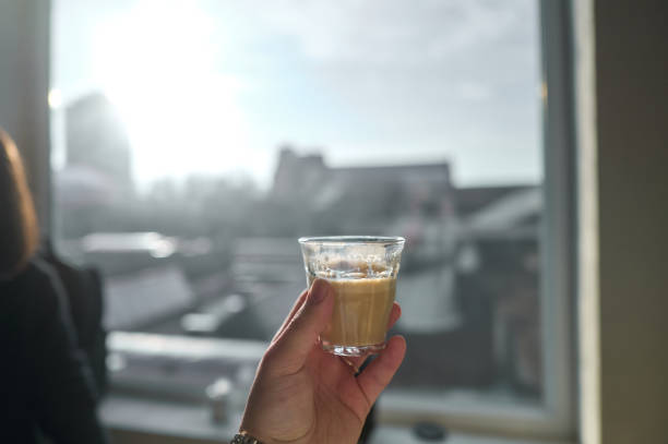 Hand holding a glass of coffee against a bright window with a blurred city view