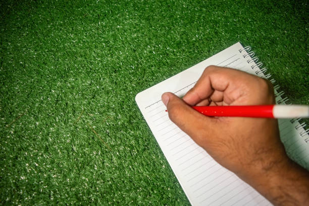 A hand holding a red pen is writing on a lined notebook placed on a green grass surface. Ideal for themes related to outdoor writing, creativity, and note taking