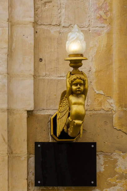 A vintage golden angel-shaped lamp ornament attached to a textured stone wall. Featuring intricate details and a warm aesthetic, this decoration harmonizes historical charm with dramatic and classical indoor accents.