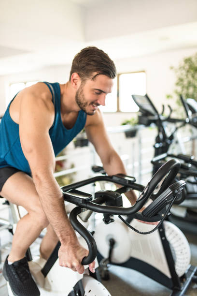 Muscular handsome Indian young man working out on exercise stationary bicycle in the gym and listening music with earphones. Health and fitness gym concept
