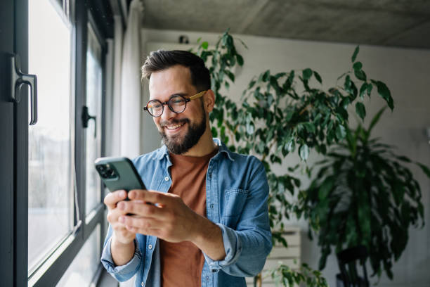 istock Young man using mobile phone and smiling near window at home 2198671499
