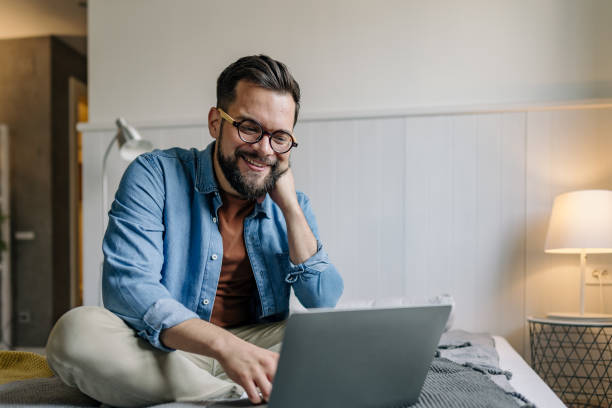 istock Smiling young man working from home sitting on bed using laptop 2198671555