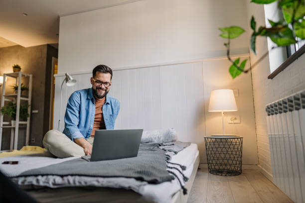 istock Young man working from home, comfortably sitting on bed using laptop 2198671568