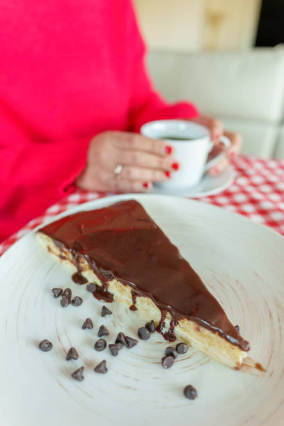 Woman with red nails is holding a cup of coffee in the background, with a slice of chocolate cake in the foreground