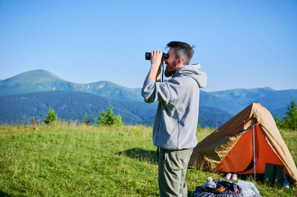 istock Man stands near orange tent, using binoculars to observe distant mountains. 2198688306