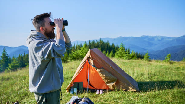 istock Man stands near orange tent, using binoculars to observe distant mountains. 2198688307
