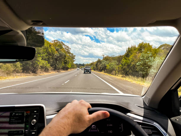 istock A wide-angle view through a car’s windscreen reveals a highway through a forested area, framed by the interior, steering wheel, and driver’s hands under a clear blue sky with fluffy clouds 2198702757