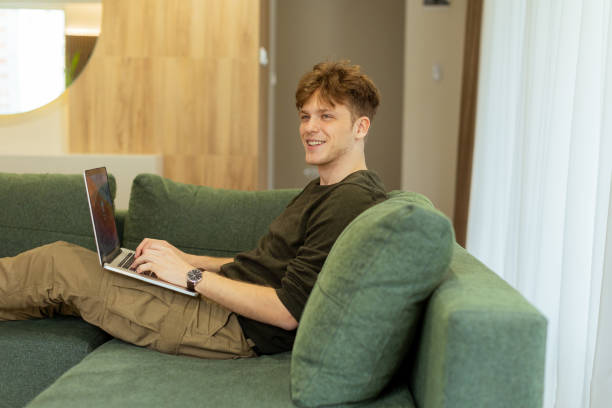 istock Man working diligently on his laptop while relaxing on a cozy green couch in a bright, airy living room 2198702907