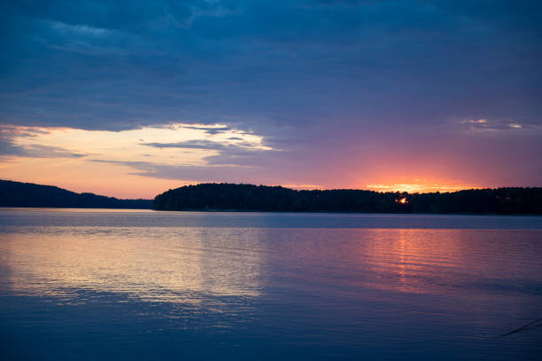 This serene view of Lake Ouachita, Arkansas, captures the calm waters reflecting the soft hues of an overcast sky. The lake stretches into the distance, surrounded by rolling, tree-covered hills and small islands. The cloud cover creates a dramatic yet peaceful atmosphere, with patches of sunlight casting a golden glow on the horizon. Gentle ripples on the water add texture, enhancing the sense of tranquility. The balance between the dark clouds and the warm light makes this a stunning depiction of nature’s beauty at dusk or dawn.