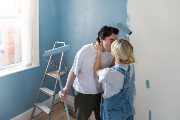 Young couple decorating their first home together, painting their walls blue. They are first time home owners in the North East of England. They're kissing each other.Videos are available similar to this scenario.