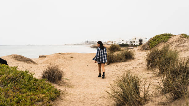 istock A young woman walks along a sandy coastal path surrounded by dunes and vegetation. 2198710941