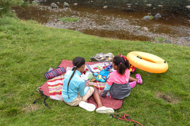 Wide shot of two sisters sitting down having a picnic by a river at Ingram Valley in the Northeast of England. They are on a family day out, sitting on blankets while playing on their mobile phones.\n\nVideos are available similar to this scenario.