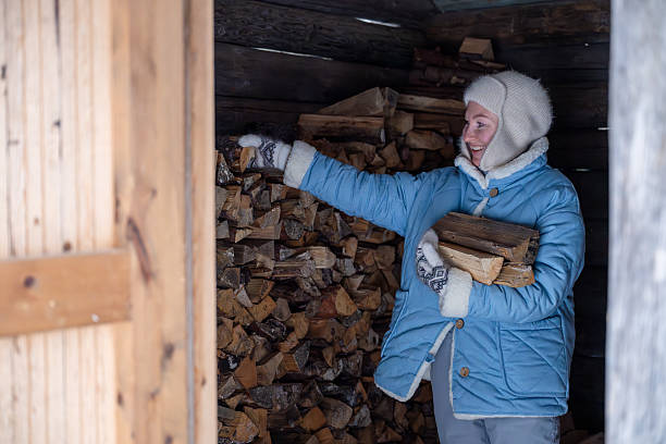 woman in winter jacket and mittens stacking firewood inside a rustic wooden shed.