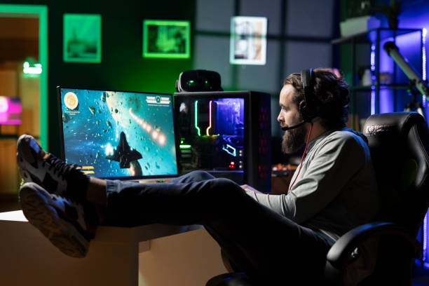 istock Man talks with friends while playing videogame with spaceship, lounging on desk 2198728426