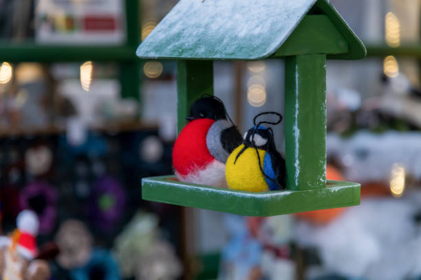Two vibrant felt toy birds in red, yellow, and black sits on a green bird feeder surrounded by a wintery market decorated with soft lights and festive items.
