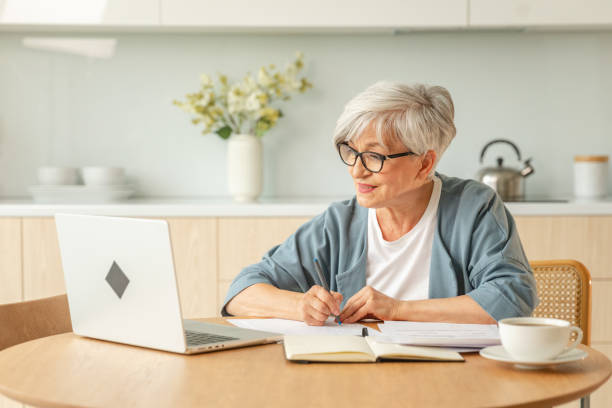istock Middle aged senior woman using laptop computer writing notes at home. Focused mature old businesswoman work in home office making written records doing online work. Mature boss professional worker. 2198760397