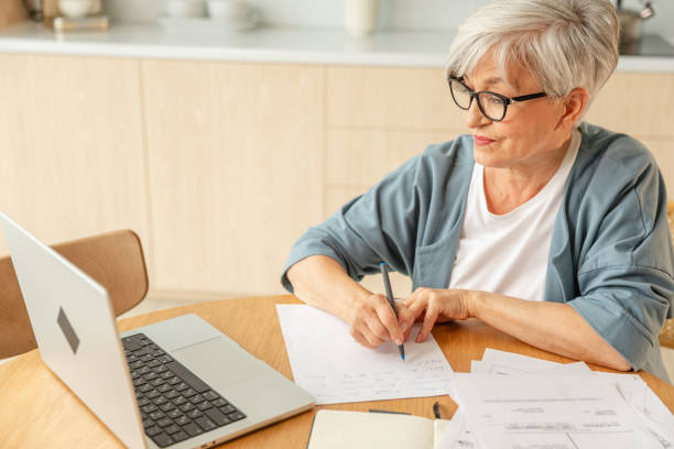 istock Middle aged senior woman using laptop computer writing notes at home. Focused mature old businesswoman work in home office making written records doing online work. Mature boss professional worker. 2198760404