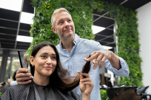 Hairdresser talks to a female client sitting in a hairdressing chair about the damaged ends of her long hair and about the haircut and new hairstyle. The hairdressing salon has green decoration with plants on the wall.