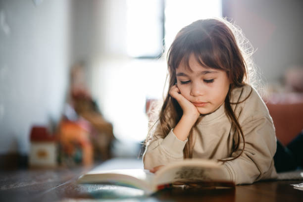 istock Beautiful little girl reading book lying on the carpet 2198765308
