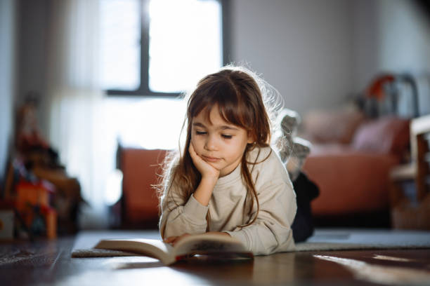 istock Beautiful little girl reading book lying on the carpet 2198765370