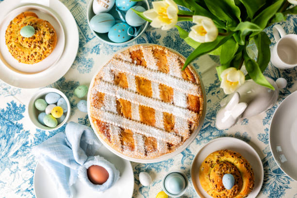 istock Top view of served easter round dining table, Italian Easter cake, shortcrust pie with ricotta cheese, Pastiera Napoletana, colorful eggs, buns, easter bunny and bird sculptures, vase with tulips flowers. Blue and white table cloth. 2198766878