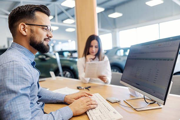 istock Smiling car seller working with paperwork on computer while lady buyer reading a contract at car showroom. 2198768927