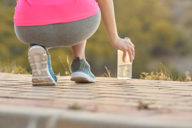 A person in athletic wear is squatting down to pick up a cristal water bottle while exercising outdoors. The scene captures a sunny day and well-maintained path in a park setting.