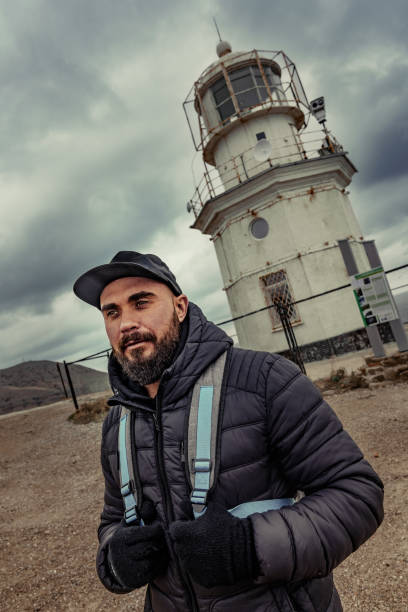 A man poses confidently in front of a weathered lighthouse surrounded by a dramatic sky. The atmosphere is moody and reflects the coastal charm of the area.
