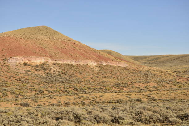 A serene landscape featuring rolling hills covered in sagebrush with distinct red rock layers exposed along the hillside. The arid terrain and earthy tones highlight the rugged beauty of the American West. Ideal for themes of geology, desert ecosystems, and remote wilderness