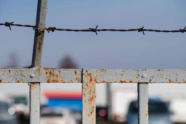 A close-up view of a rusty metal fence topped with barbed wire, set against a blurred urban backdrop. The contrast between the weathered texture of the fence and the soft background creates a striking urban composition.