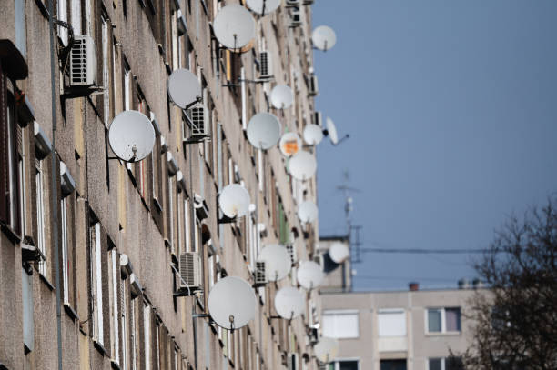 The facade of an apartment building is covered with numerous satellite dishes, showcasing modern urban living and communication infrastructure. The clear blue sky contrasts with the busy arrangement of dishes and windows.