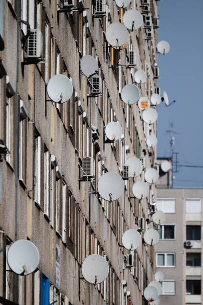 The facade of an apartment building is covered with numerous satellite dishes, showcasing modern urban living and communication infrastructure. The clear blue sky contrasts with the busy arrangement of dishes and windows.