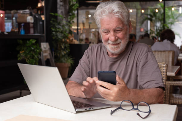 istock Attractive senior bearded man sitting in a cafe using laptop and mobile phone while enjoying a break. Modern and smiling elderly man working in a relaxed atmosphere 2198839040
