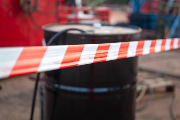 Red-white tape line for barrier hazard area with chemical containment as blurred background. Industrial unsafe workplace, close-up at foreground.