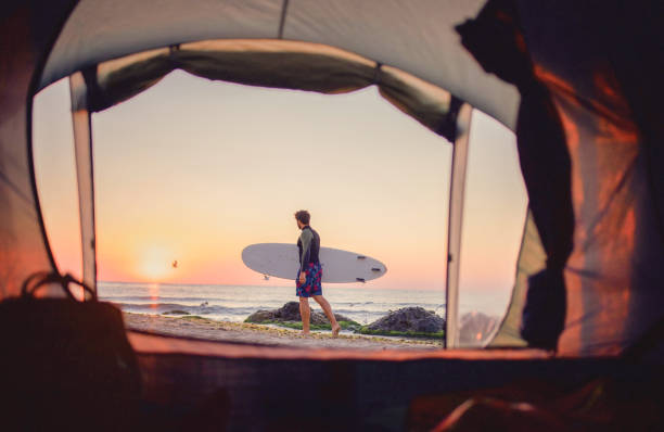 View from camping tent on full length surfer walking on the beach at sunrise.