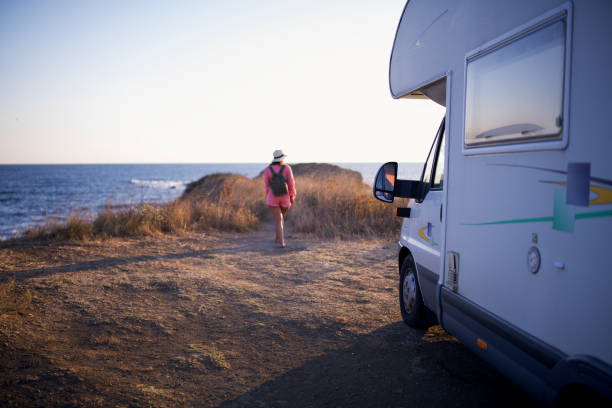 Young woman with a straw hat and backpack is watching the view of the sea and walking next to her camper