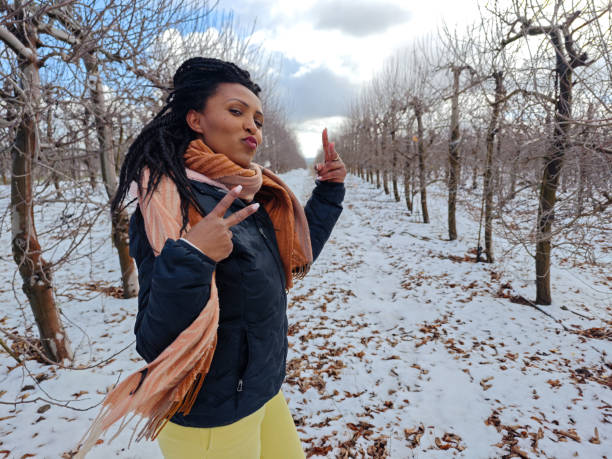 A stylish woman stands in a snow-covered orchard, playfully making a peace sign while pouting at the camera. She is dressed in a dark blue winter jacket, a warm beige scarf, and yellow pants, exuding confidence and fun energy. The winter landscape with leafless trees and a soft cloudy sky creates a serene backdrop, contrasting with her vibrant and charismatic expression. This image captures a joyful, carefree moment filled with personality and winter charm.