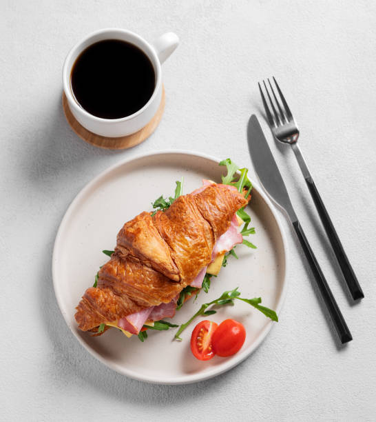 Croissant sandwich with ham, cheese, tomato  and arugula on a plate on a light background