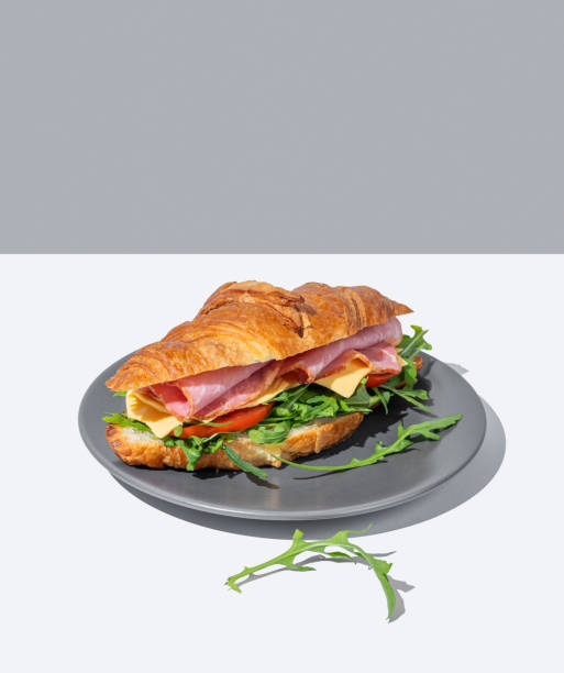 Croissant sandwich with ham, cheese and arugula on a dark plate on a light background with a shadow.