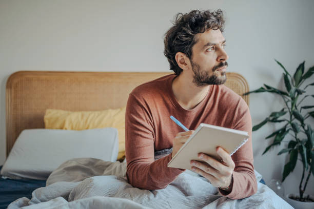 Thoughtful Young Handsome middle eastern man sitting in bed and journaling or writing in his notebook. Morning Creativity & Reflection.