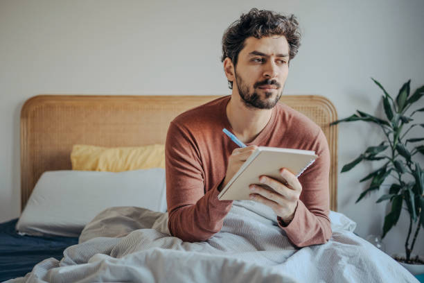 istock Thoughtful Young Handsome middle eastern man sitting in bed and journaling or writing in his notebook. Morning Creativity & Reflection. 2198868814