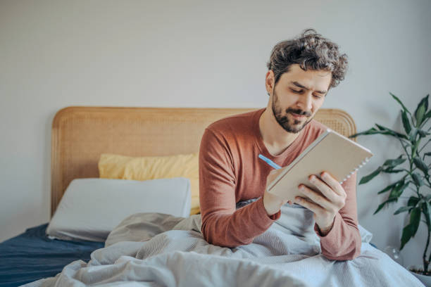 istock Thoughtful Young Handsome middle eastern man sitting in bed and journaling or writing in his notebook. Morning Creativity & Reflection. 2198868841