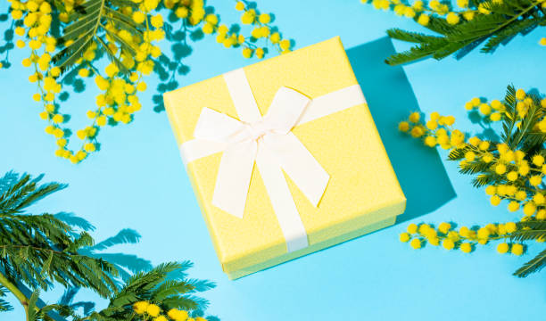 istock Yellow gift box on blue background with mimosa flowers and sunlight. 2198869850
