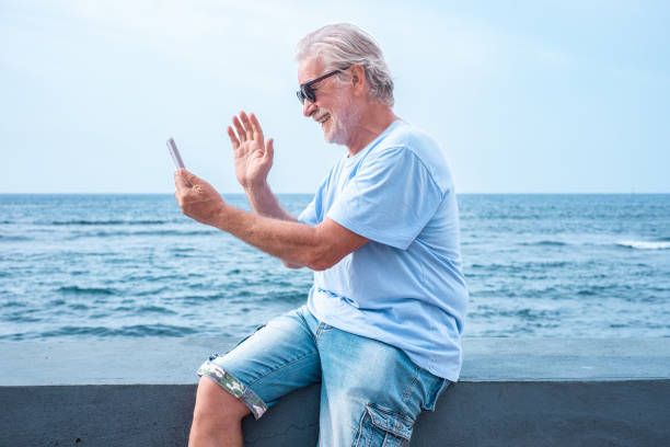 istock Cheerful senior man with beard sitting on sea shore using cellphone for video call, elderly pensioner enjoying freedom, vacation or retirement. Horizon over water 2198872384