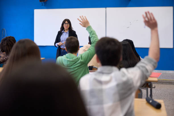 A young ethnic female professor stands at a large whiteboard and calls on a student who is raising his hand. They are learning about conjugating Spanish verbs. The shot is from the back of the classroom.
