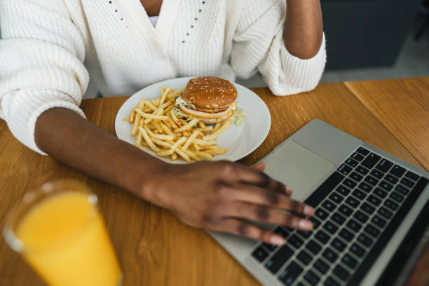 Woman is working on her laptop while enjoying a tasty lunch of burger and fries, accompanied by a refreshing glass of orange juice, showcasing the concept of working from home and multitasking
