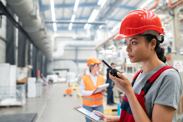 istock Female asian engineer wearing hard hat and using walkie talkie while working on industrial factory 2198886298