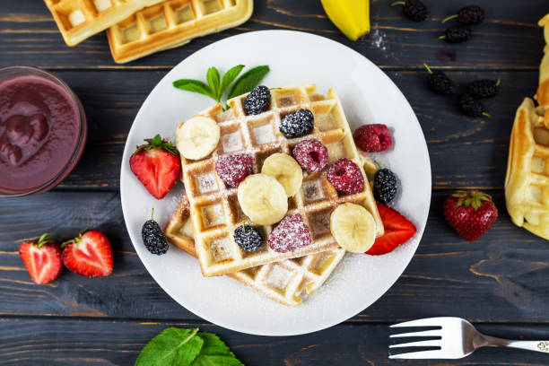 istock Square Belgian waffles with berries and powdered sugar on a white plate on a wooden table. Delicious sweet sweet waffles. View from above. 2198892462